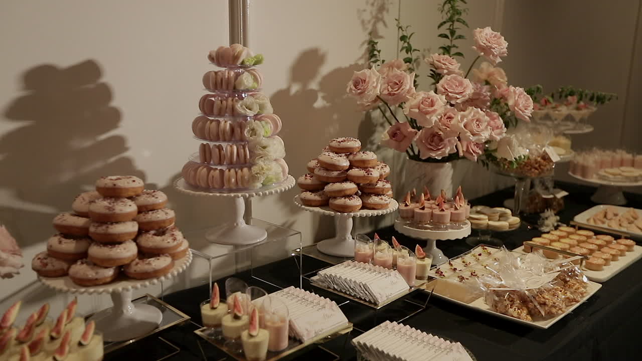 Kameraet panorerer og viser et buffetbord med lækre desserter i et lyserødt dekorationstema, med smukke blomsterarrangementer