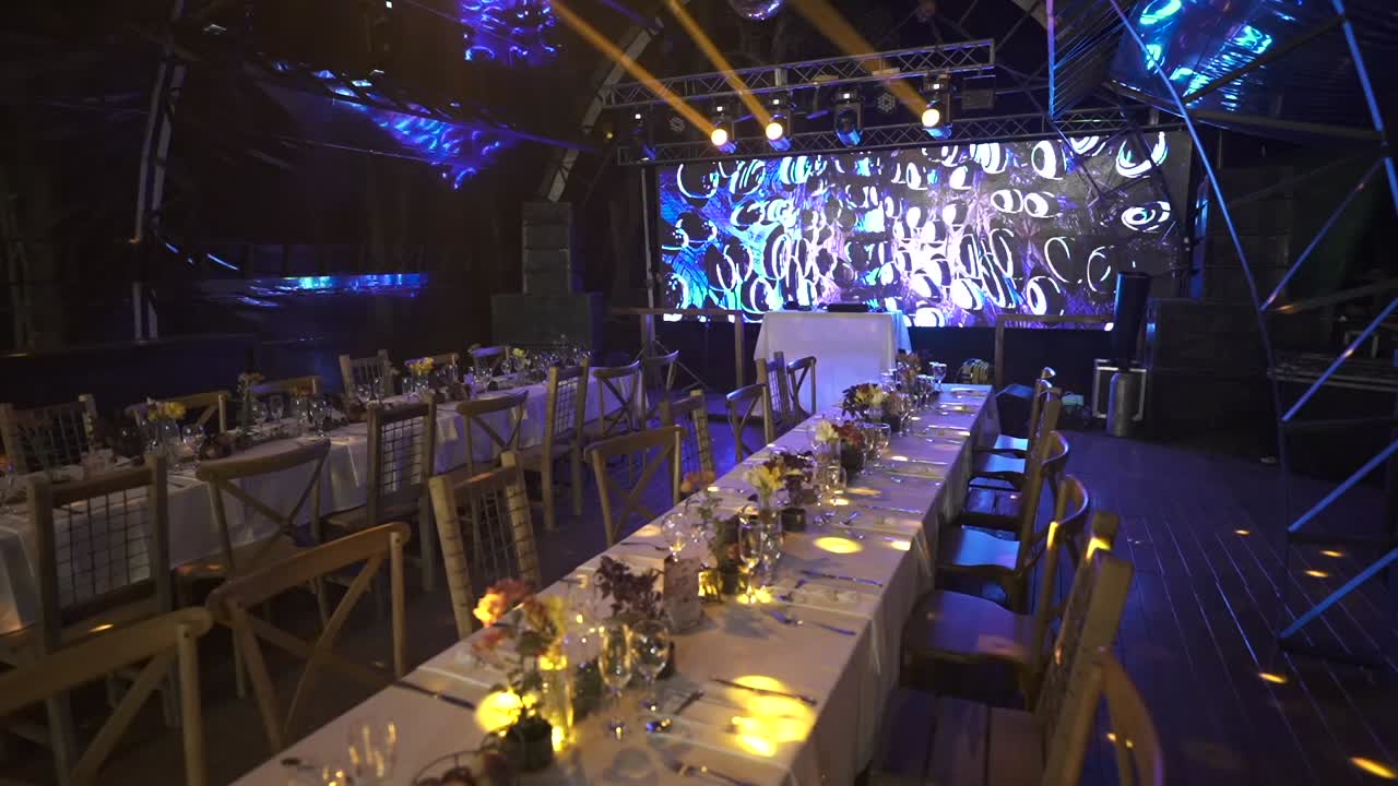 Wide, empty event hall features long banquet settings, detailed table decor, staged blue and gold lighting, and modern AV setup, ready for a formal celebration