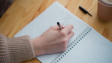 close-up of young student hand writing with pen in notebook, partial view of coffee cup on table, demonstrating a focused study moment with soft background