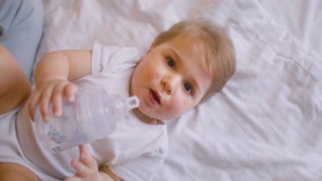 vista superior de un bebé acostado en la cama y mirando la cámara en el dormitorio mientras bebe agua del biberón