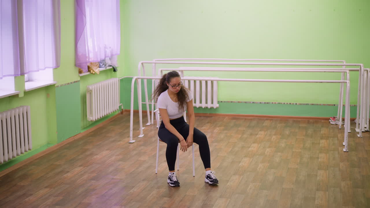 Close view of girl in white top and black leggings with canvas sneakers sitting on chair inside studio, looking stressed and exhausted after workout, wooden floor and mirror reflection in background