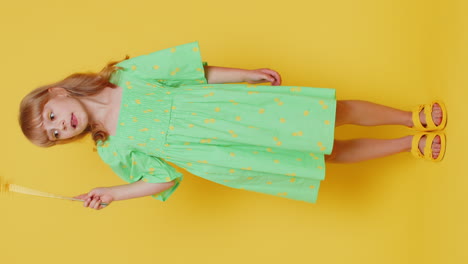 a young girl in a green dress smiles while holding a star-shaped wand on a yellow background.