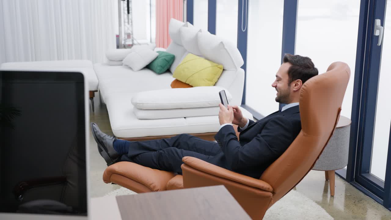 Happy smiling man in a suit relaxes in a modern leather chair while using his smartphone in stylish living room with colorful cushions and large windows. Businessman resting in office watching phone