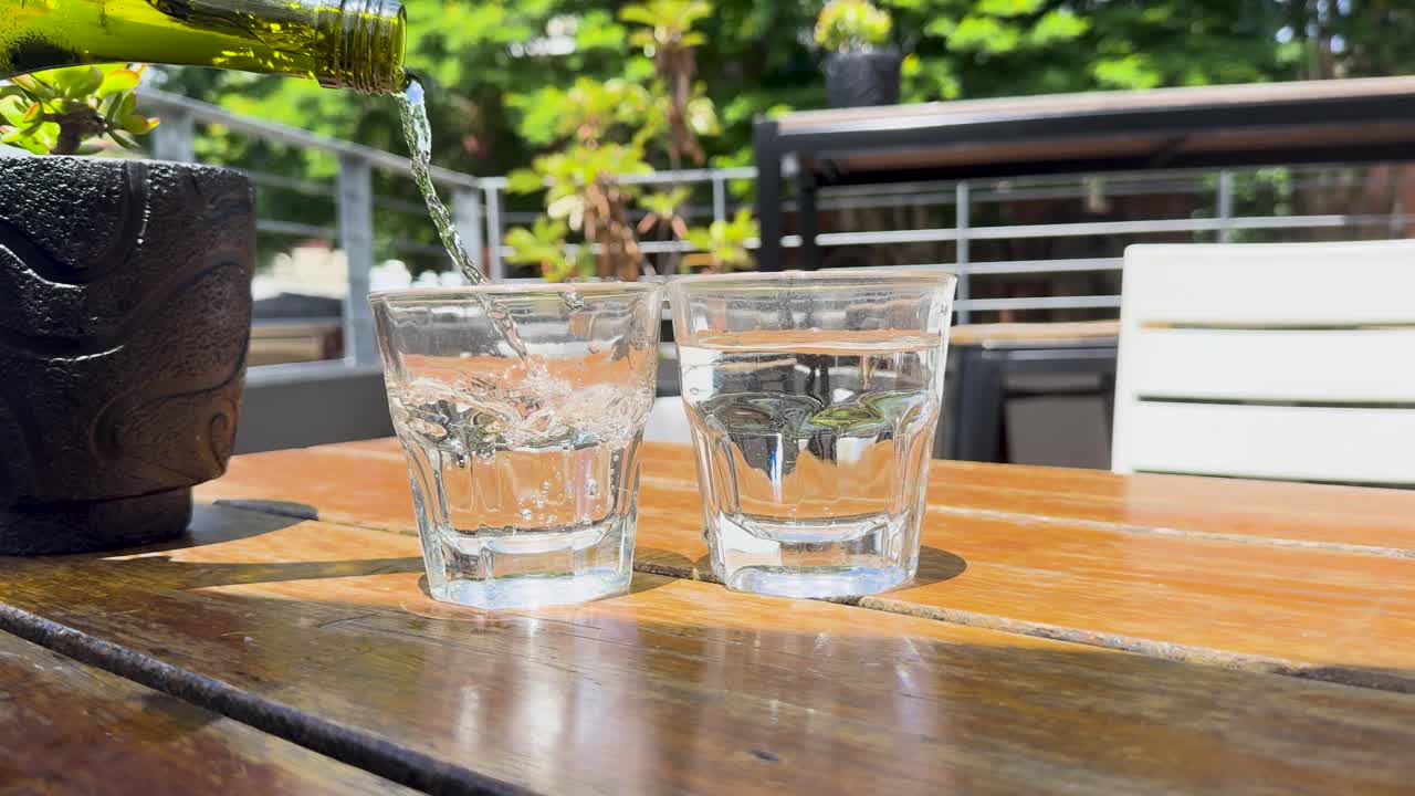Water is poured into glasses on a sunlit outdoor table, creating a refreshing scene with vibrant greenery in the background