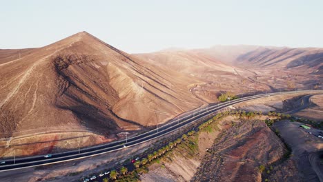 Camino-Sinuoso-A-Través-De-Colinas-áridas-En-Fuerteventura,-Con-Escasa-Vegetación-Y-Autos-Estacionados,-Vista-Aérea