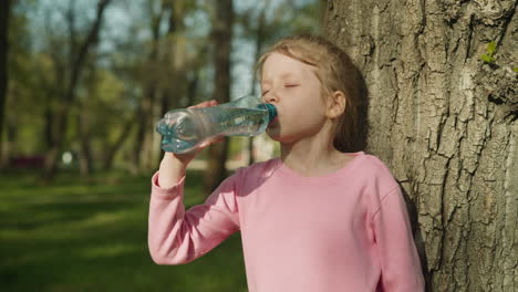 una chica rubia sedienta bebe agua cerca de un árbol viejo en el parque de la ciudad