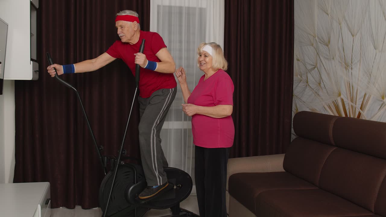 활동적인 성숙한 노인 할아버지 스포츠 의류를 입은 남자가 오비트렉을 사용하고, 할머니는 집에서 그를 격려합니다.
