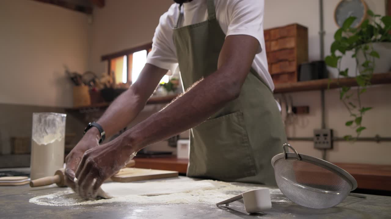 African man in green apron pressing and rolling dough on counter, making pastry with rolling pin