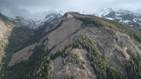 Side-panning-shot-of-summer-mountains-at-low-altitude-and-snowy-cloudy-peaks-near-the-top,-shot-in-Big-Cottonwood-Canyon,-Utah-with-sunset-lighting-coming-in-from-the-right-side