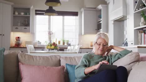 happy senior caucasian woman sitting on sofa in living room, using tablet