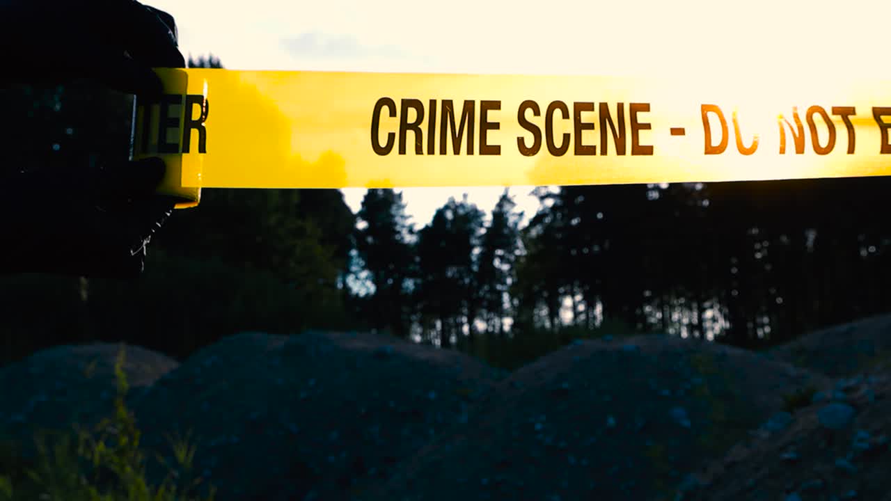Close up view of a police investigator with black nylon forensic gloves unravel or unrolling a yellow crime scene tape in front of a nature area where large rocky piles of gravel are at sunset night