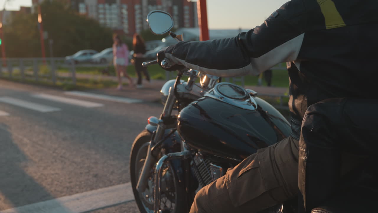 Biker waits on motorcycle at crosswalk while pedestrian carrying scooter passes across street in warm evening sunlight reflecting on asphalt creating cinematic urban transport