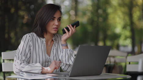 Mujer-De-Negocios-Segura-Grabando-Mensajes-De-Audio-En-Un-Teléfono-Inteligente-En-La-Calle.-Una-Mujer-Sentada-En-Un-Café-De-Verano-Utiliza-El-Reconocimiento-De-Voz-De-Un-Teléfono-Inteligente-Para-Enviar-Mensajes-De-Voz.-Concepto-De-Altavoz-O-Mensaje-De-Voz-Saliente