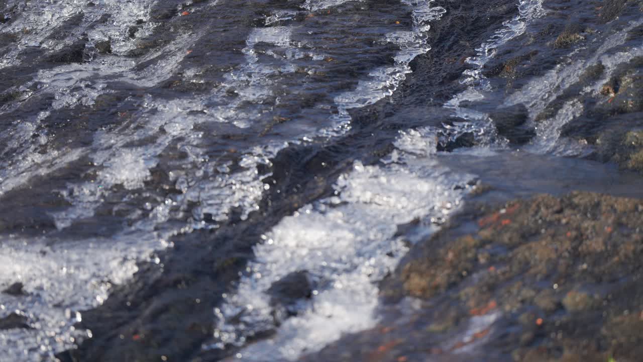 Un piccolo rivolo di acqua di fusione scorre sulla filigrana di ghiaccio sottile sulle rocce scure.