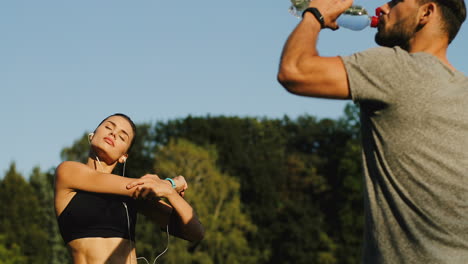 Rückansicht-Des-Trinkwassers-Des-Joggers-Und-Der-Jungen-Frau-Mit-Kopfhörern,-Die-An-Einem-Sonnigen-Sommertag-Hände-Und-Schultern-Trainieren-Und-Aufwärmen