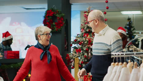 cheerful elderly couple dancing in christmas decorated clothing store, feeling xmas holiday mood spirit while browsing for presents. senior customer being spinned by husband in fashion boutique