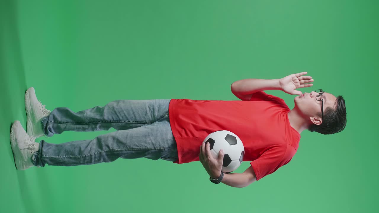 공을 들고 있는 아시아인 남성의 몸이 가득 차 있으며, 초록색 스크린 배경에서 축구를 응원하는 동안 패배자들을 조롱하고 있다.