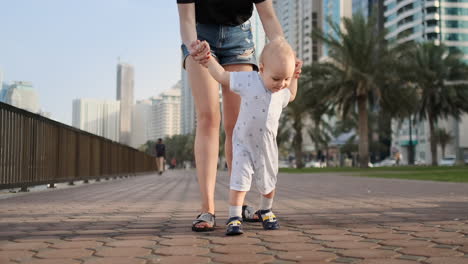 Madre-Joven-Con-Un-Niño-Y-Primeros-Pasos.-Madre-Joven-Con-Un-Niño-En-El-Aprendizaje-Al-Aire-Libre-Para-Dar-Los-Primeros-Pasos-Cerca-De-La-Ciudad-Urbana