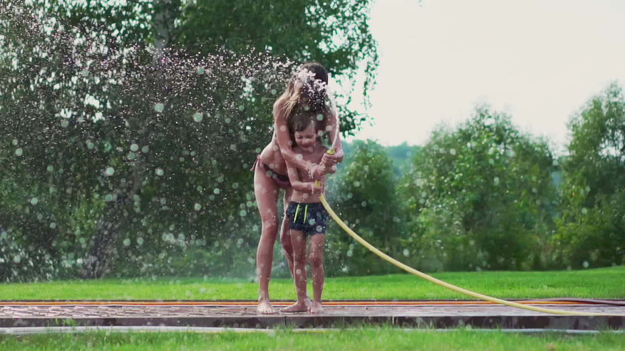 Mom and son playing on the lawn pouring water laughing and having fun on the Playground with a lawn on the background of his house near the lake
