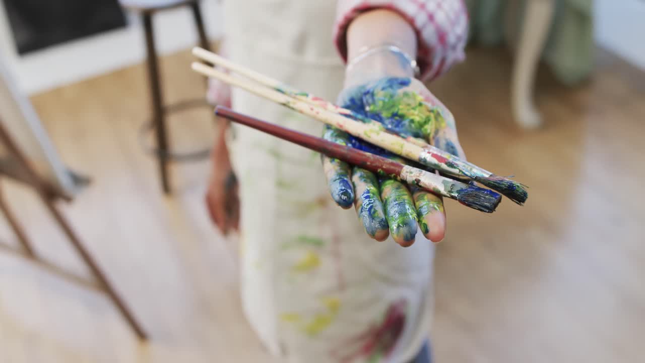 Mano di un'artista donna birazziale con grembiule che tiene pennelli da pittura in uno studio d'arte, in movimento lento.