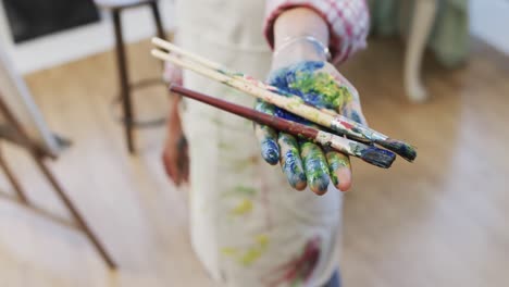 hand of biracial female artist in apron holding paint brushes in art studio, slow motion