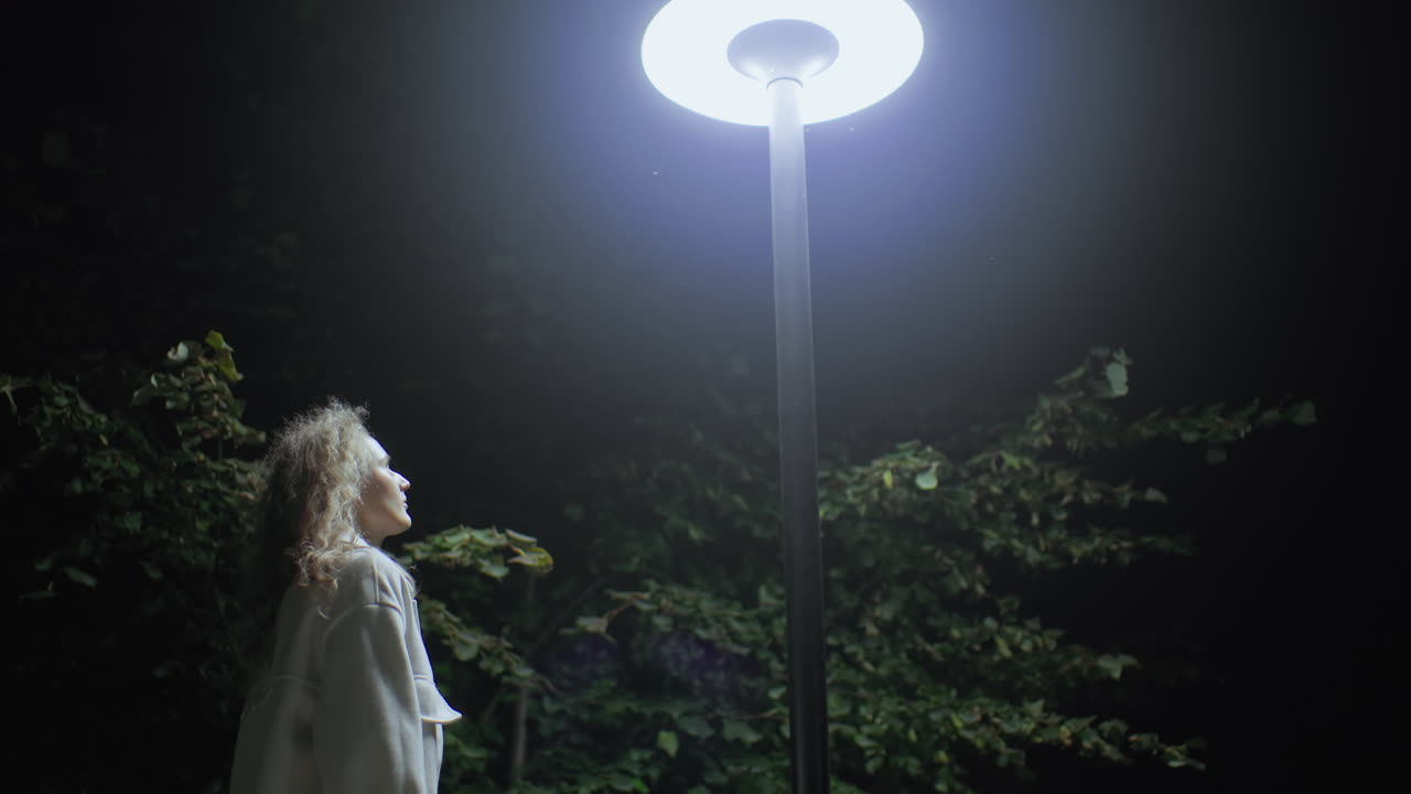 Girl in white coat stands, gazes upward at bright lamp amid leafy branches, nighttime setting creating soft glow over her contemplative expression