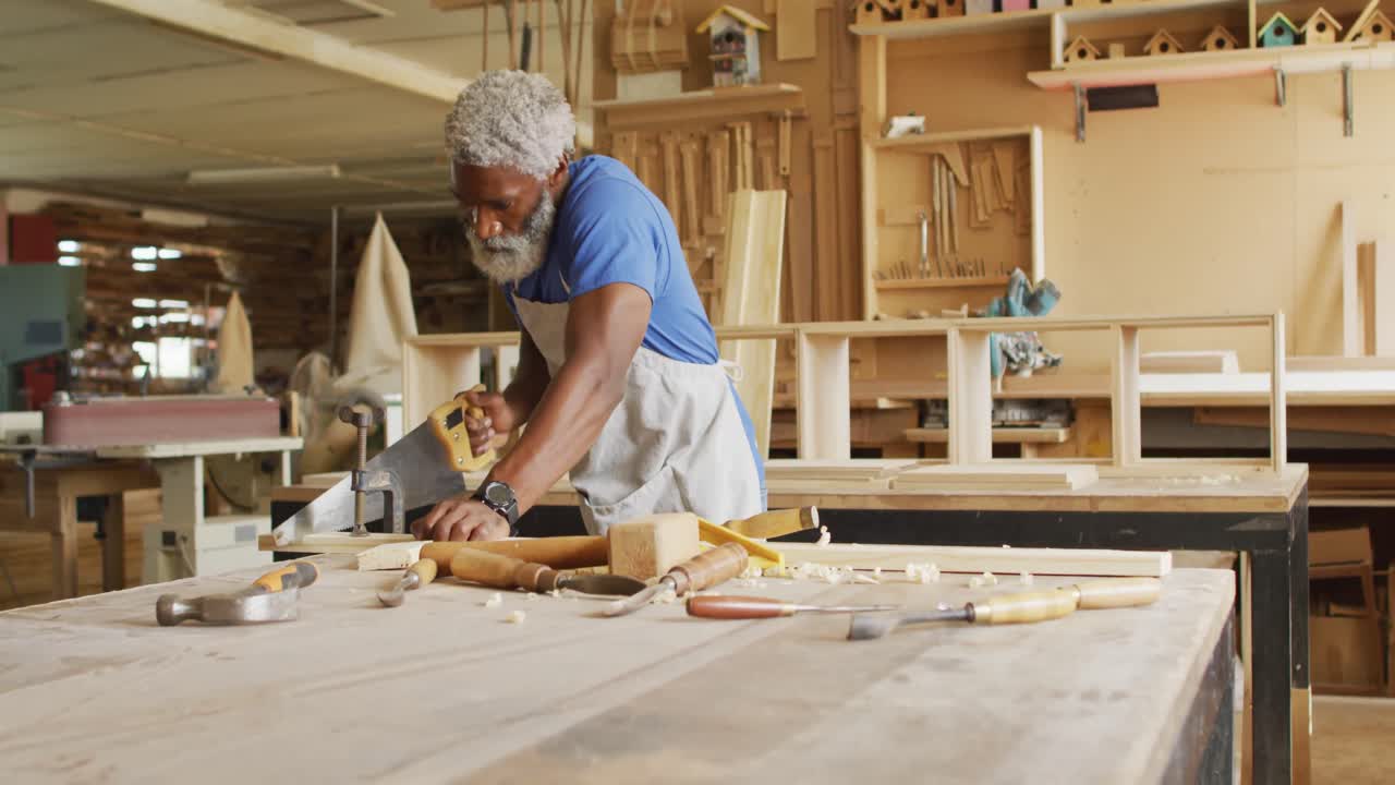 African american male carpenter cutting a wooden plank using a hand saw in a carpentry shop