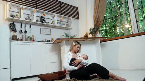 blonde woman sits on the floor covered with plaid in the kitchen. mother waving her baby and giving a pacifier.