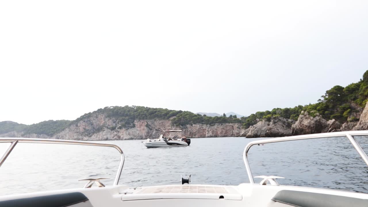 The bow of a speedboat sailing through crystal clear water in Croatia. The cliffs of Kolocep Island with other boats moored