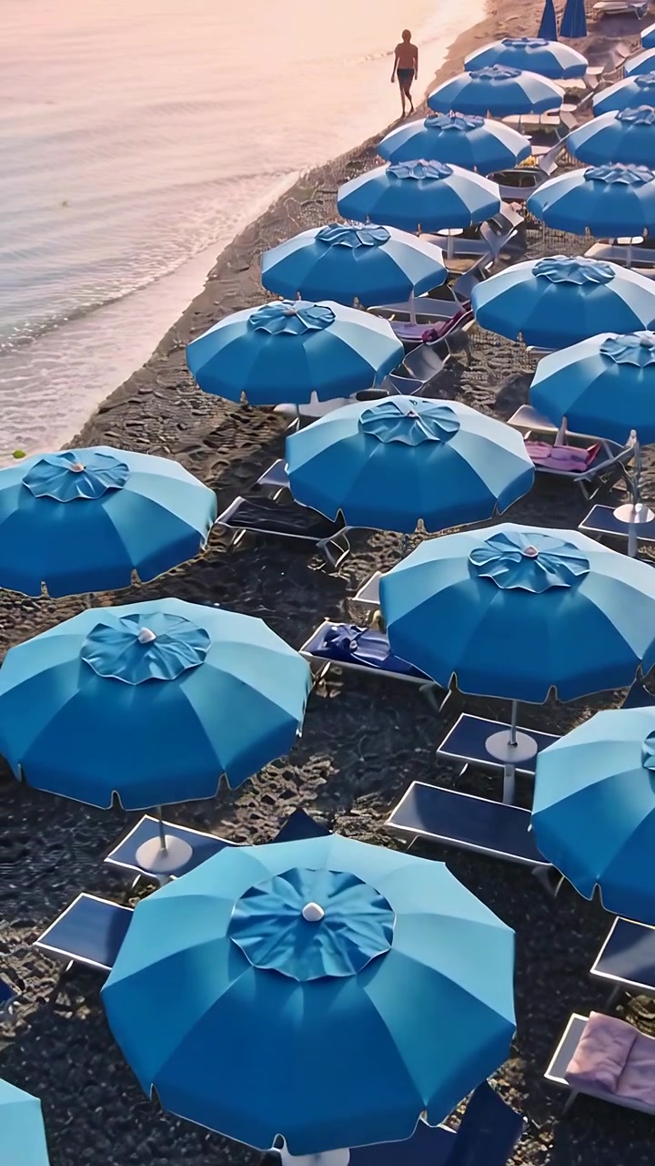 Video verticale: Uomo in costume da bagno che arriva, camminando su una spiaggia di ciottoli per una passeggiata tranquilla con ombrelloni blu.