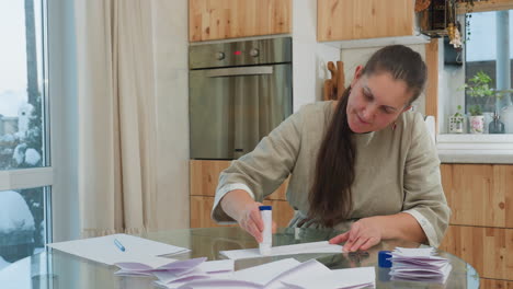 woman applying glue to paper while sitting at table with scissors, folded paper, and other crafting supplies in background