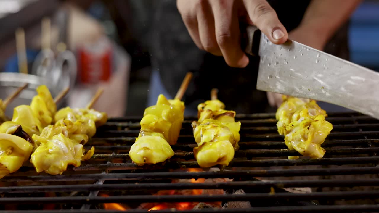 Street vendor grills yellow-marinated squid skewers over open flame, using knife, close-up view
