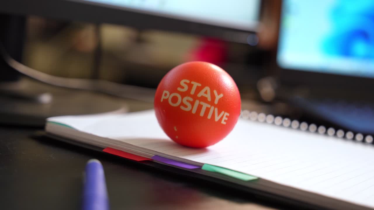Orange stress ball with "STAY POSITIVE" text on a notebook in office setting. Perfect for motivation, workplace, or mental health themes.