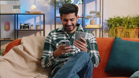 Smiling-happy-indian-man-counting-money-cash-use-smartphone-income-saves-lottery-win,-budget-at-home