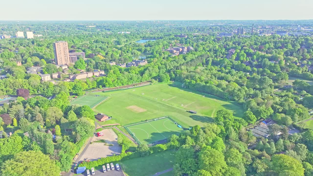 Slow glide drone over Birmingham Botanical Gardens, tall University tower building beside large sport fields surrounded by lush greenery, bright daylight highlighting urban-natural blend