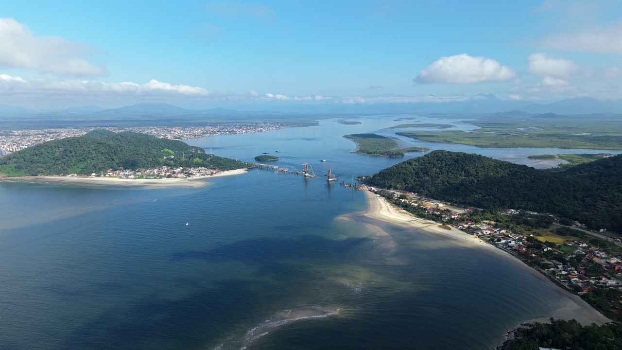 Drone glides high over Baía de Guaratuba showing bridge works connecting Guaratuba and Matinhos, with sandbanks, estuary, coastal towns, lush hills, and Serra do Mar in the distance under clear skies