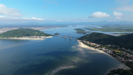 drone glides high over baía de guaratuba showing bridge works connecting guaratuba and matinhos, with sandbanks, estuary, coastal towns, lush hills, and serra do mar in the distance under clear skies