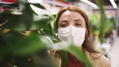 beautiful girl wearing protective medical mask and fashionable clothes looks at camera behind of plants