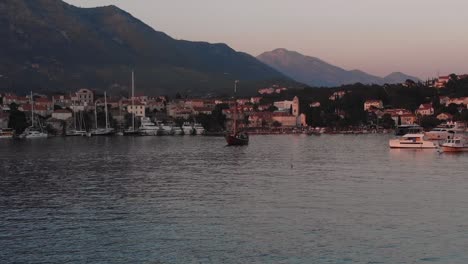 Aerial-reveal-sliding-shot-from-walkway-to-reveal-Cavtat-harbor,-Croatia-with-luxury-boats-moored-and-the-sunset-lighting-up-the-town