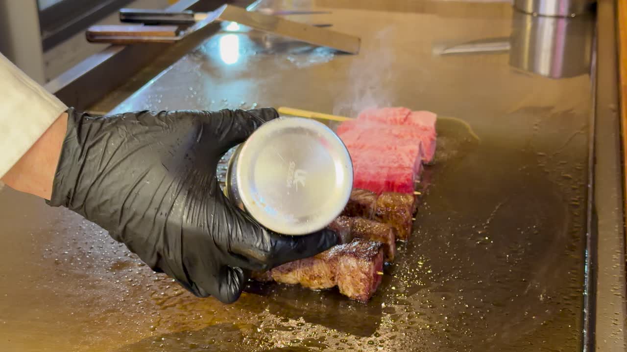黒い手袋のシェフが、熱い鉄板の上で霜降り牛串に味付け。