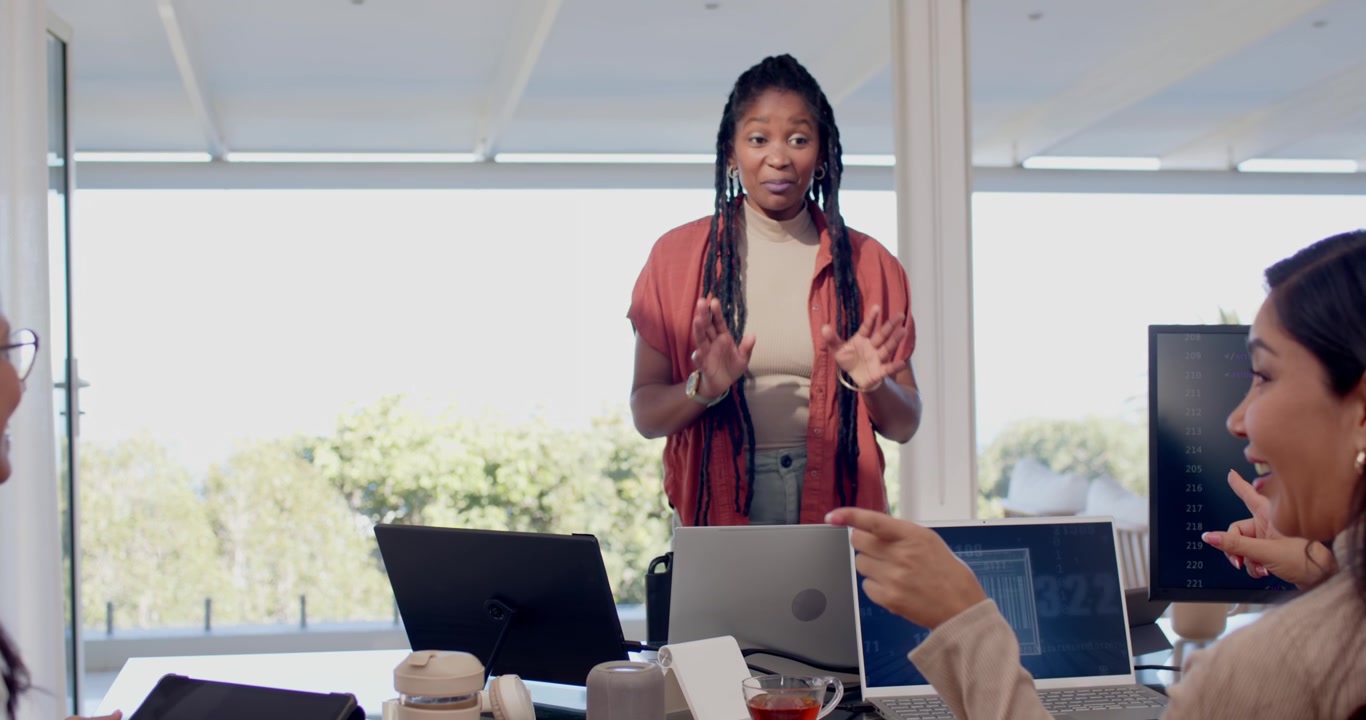Diverse female team watching presenter in rust shawl leaning over laptops leading meeting at table