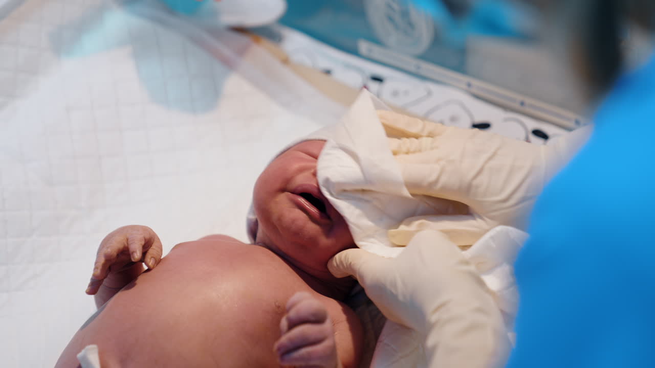 Medic's hands in latex gloves clean the face of newborn with a napkin. The doctor cutting the umbilical cord.