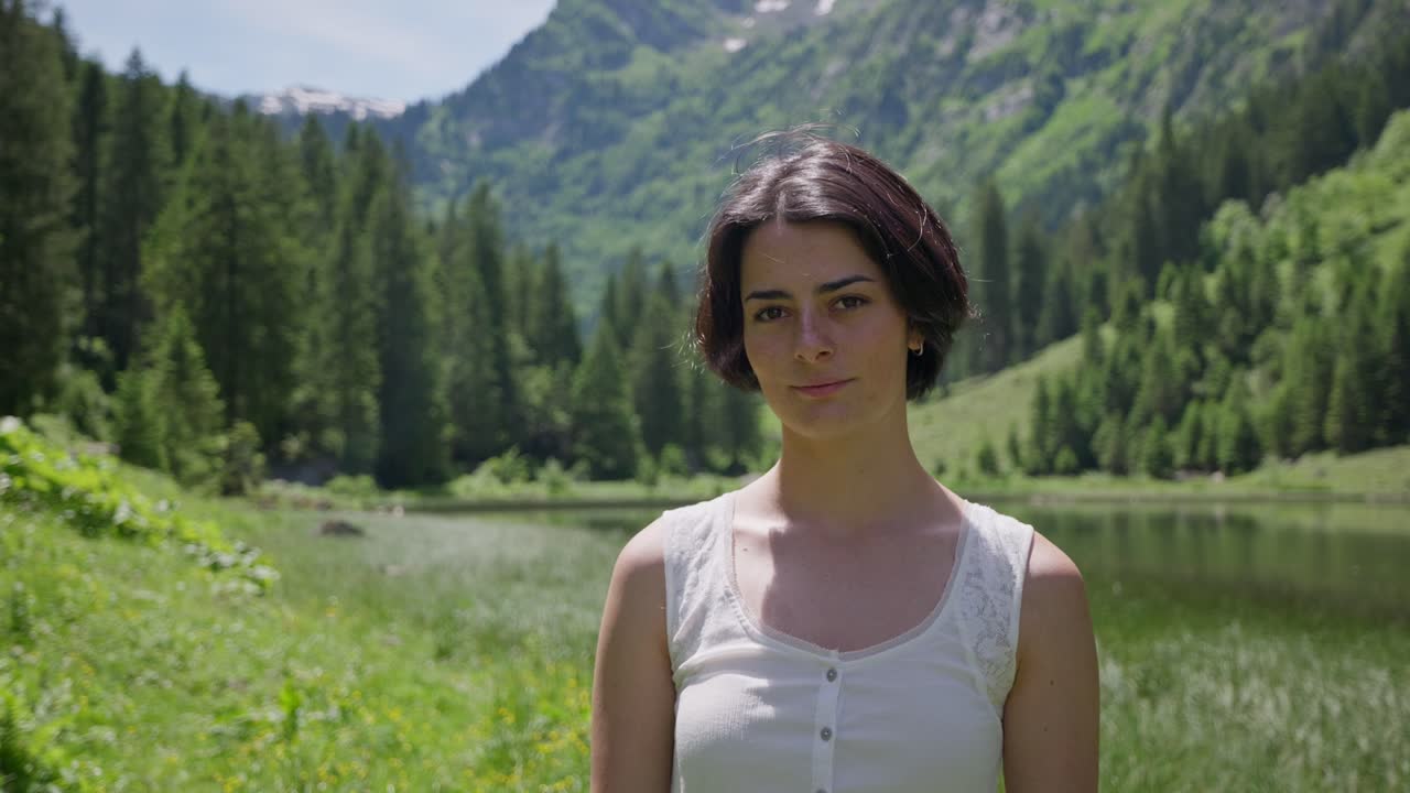 Portræt af brunette kvinde, der kigger ind i kameraet. Solrig dag udendørs foran en sø og bjerge. Nærbillede.