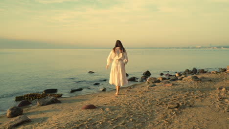 Mujer-Caucásica-Con-Un-Hermoso-Vestido-Blanco-Caminando-En-La-Playa-De-Arena-Al-Atardecer-Dorado---Turista-Femenina-En-Vacaciones-De-Verano---Seguimiento-En-Cámara-Lenta