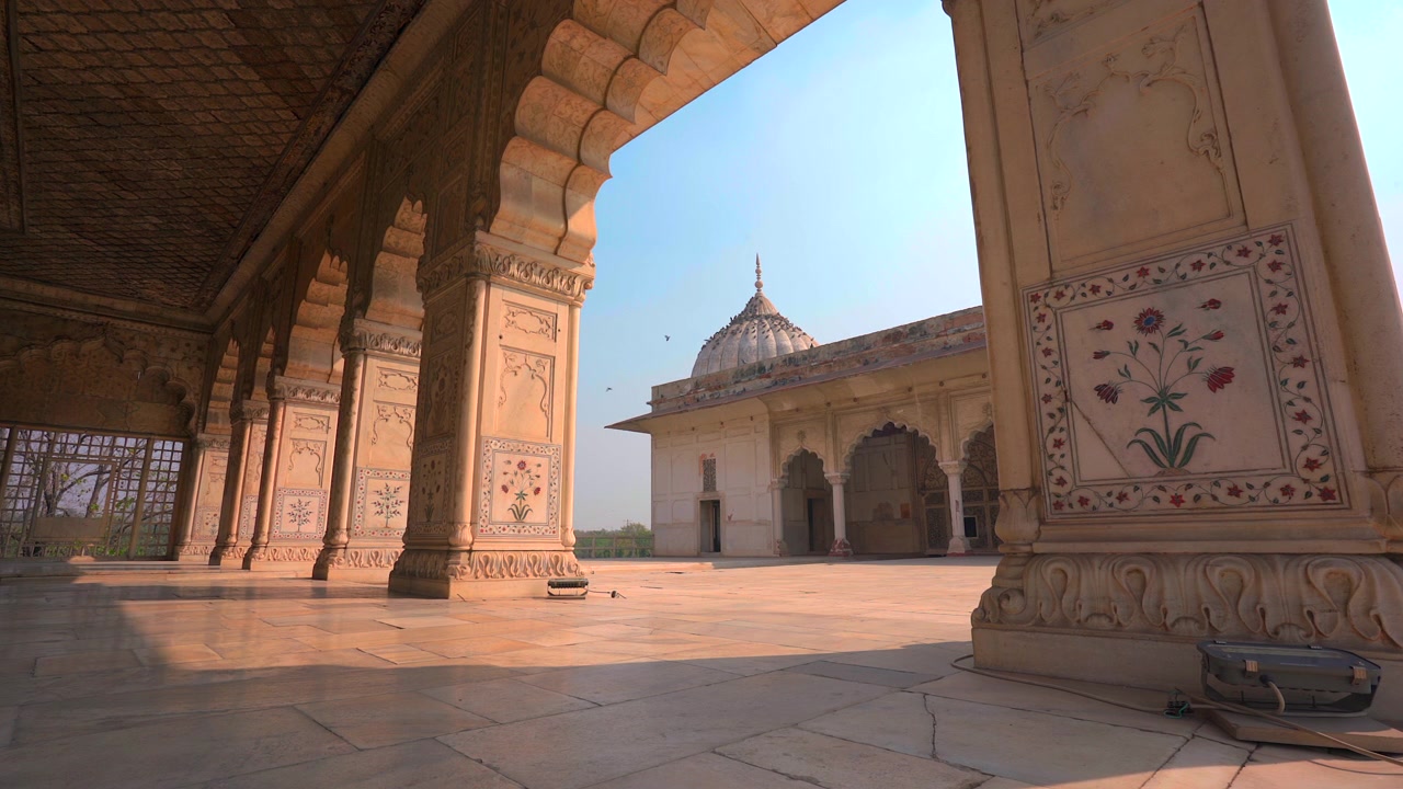 Una ripresa statica dal basso dall'interno di un padiglione intagliato in modo intricato, che inquadra una bellissima struttura a cupola in marmo bianco sotto un cielo sereno, presso lo storico Forte Rosso a Delhi, India.