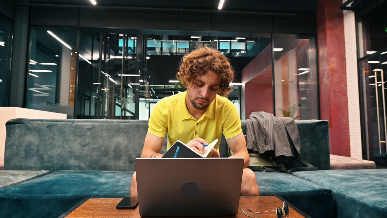 Man in yellow shirt standing on a couch and writing in a notebook and working on a computer