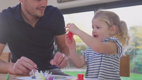 Father-and-young-daughter-having-fun-at-home-sitting-at-table-and-painting-decoration-together---shot-in-slow-motion