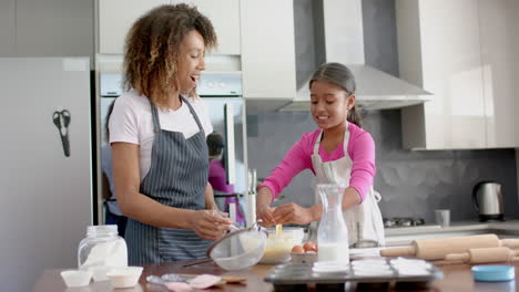 happy biracial mother and daughter making cake mix, baking in kitchen, slow motion