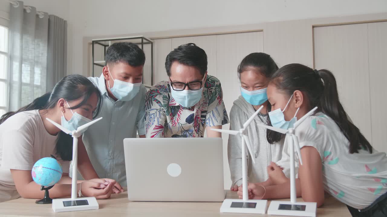 Wearing Facemasks, Enthusiastic Asian Teacher Explains To Young Children How Wind Turbines Work. Kids Learning About Eco-Friendly Forms Of Renewable Energy
