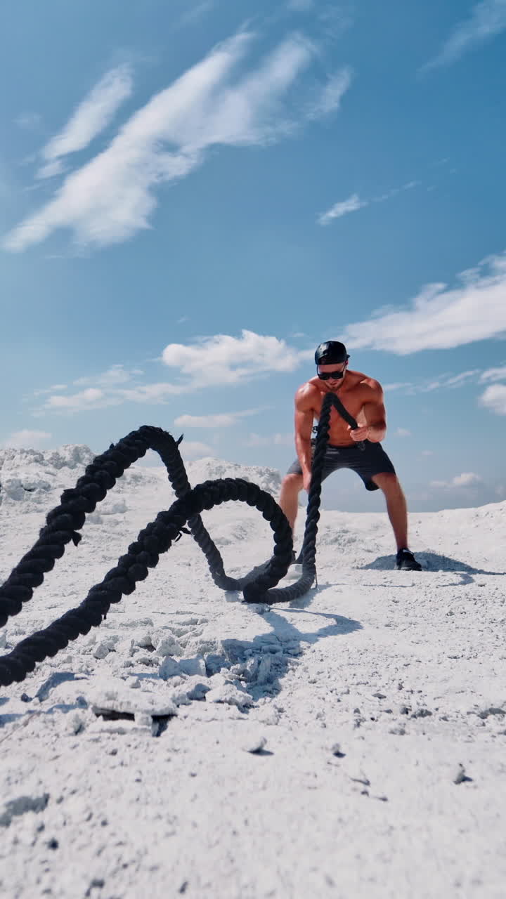 Topless man working out with a rope on nature. Muscular athlete exercising with battle ropes on the white mountain against blue sky in summer. High intensive workout. Vertical video